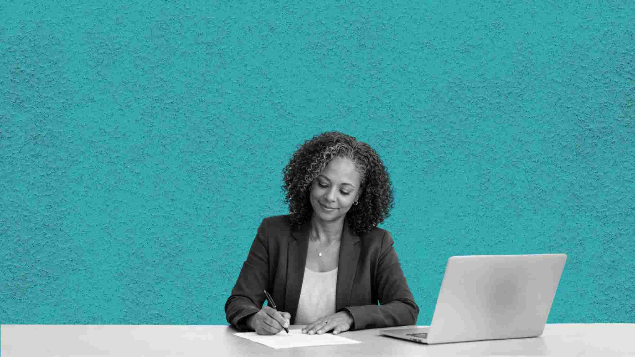 A happy female lawyer writing a letter with pen and paper next to her laptop.